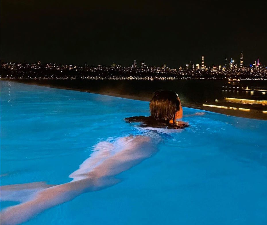 Image at night of the NYC skyline from the ange of a woman in the outdoor pool overlooking the city