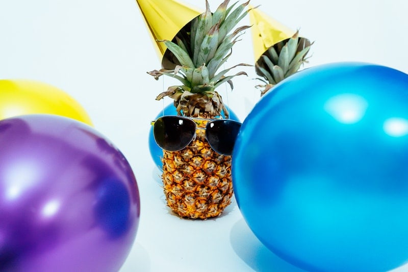 Photo of a pineapple wearing sunglasses and a party hat with balloons floating around for a birthday party