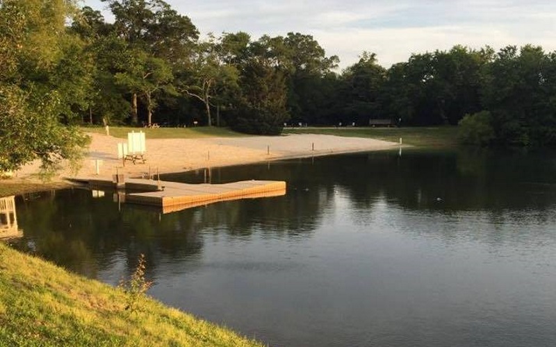 Photo of lake with a small sandy beach and  a wooden floating dock going out about 20 feet into the water