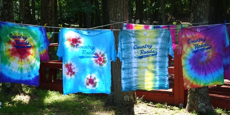 Image of four tye die tee shirts with Country Roads Day Camps logo on the front hanging on a clothes line.