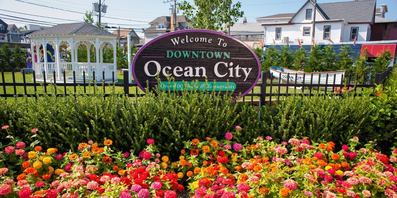 Image a sign for Downtown Ocean City, NJ with flowers blooming under it on a bright sunny day.