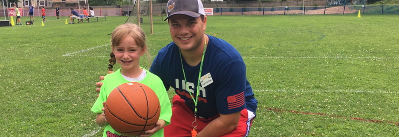 Image of a girl holding a basketball standing on a soccerfield with one of the staff at Fundamentals Day Camp.