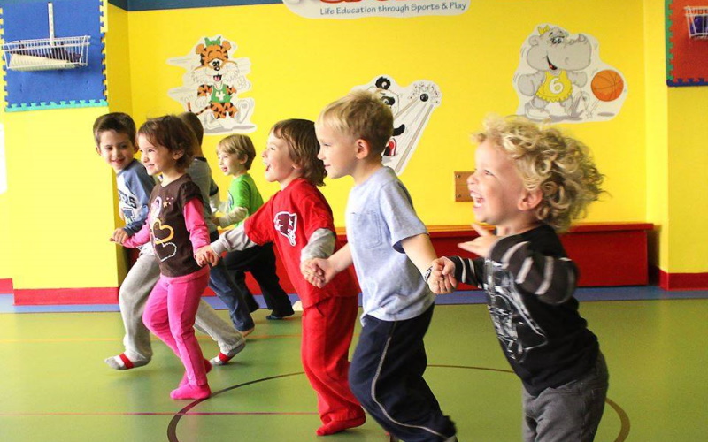 Image of seven kids holding hands playing against a yellow wall with a cartoon tiger and a cartooon rhinoceros on the wall at The Little Sport Play place in Central New Jersey