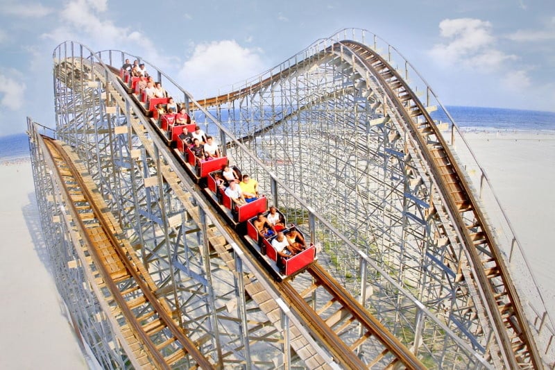 Photo of one of the best roller coasters on a beach in NJ which is the Great White wooden roller coaster with 6 red cars full of people with the beach in the background at More's Piers amusement park in Wildwood NJ