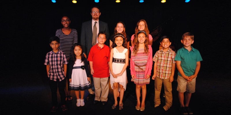 Image of a number of children standing on stage with a black background and a theater teacher next to them at MTC day camp in Marlton, New Jersey.