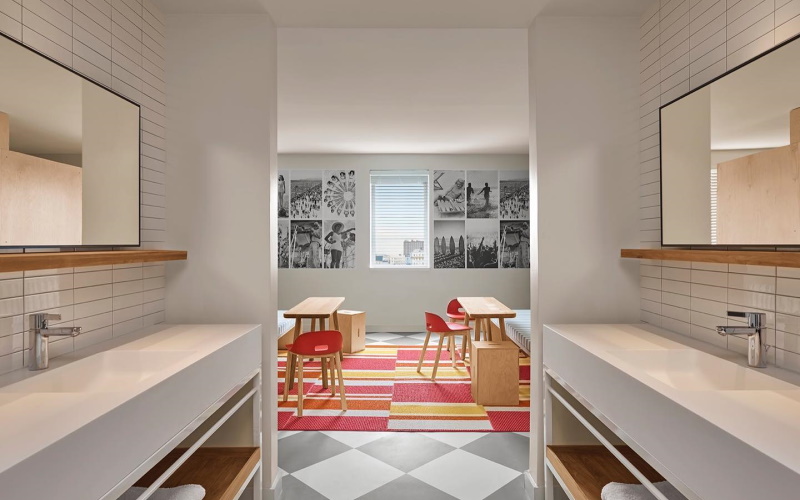 Photo of one of the rooms at the Asbury Hotel with various art hung on the wall and modern dual sink hallway and a white and black checkered floor covered partially by a red and yellow rug.