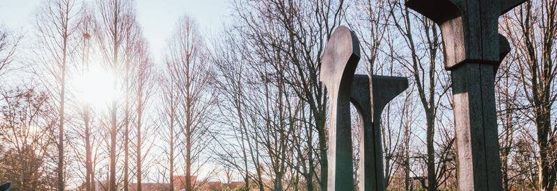 Photo of a sculpture with the sun shining behind it through some wooded bare trees at the Grounds for Sculture in Hamilton NJ