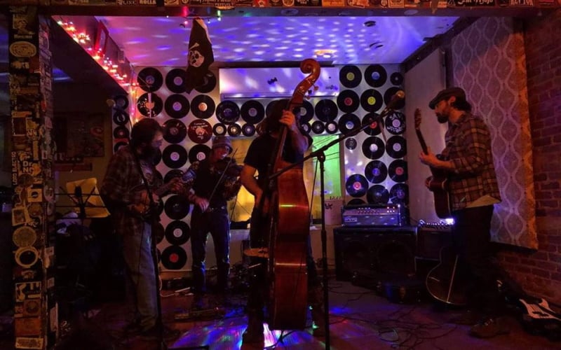 Photo of a band playing at The Saint in Asbury with a stand up bass in front and records hanging on the wall with colorful lights illuminating the wall.