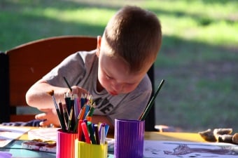 Image of a kid sitting outside at a table using a paintbrush to paint on a canvas