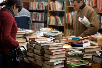 Image of books piled up on a table with multiple bookshelves in the background showing a bookstore for shopping in NJ